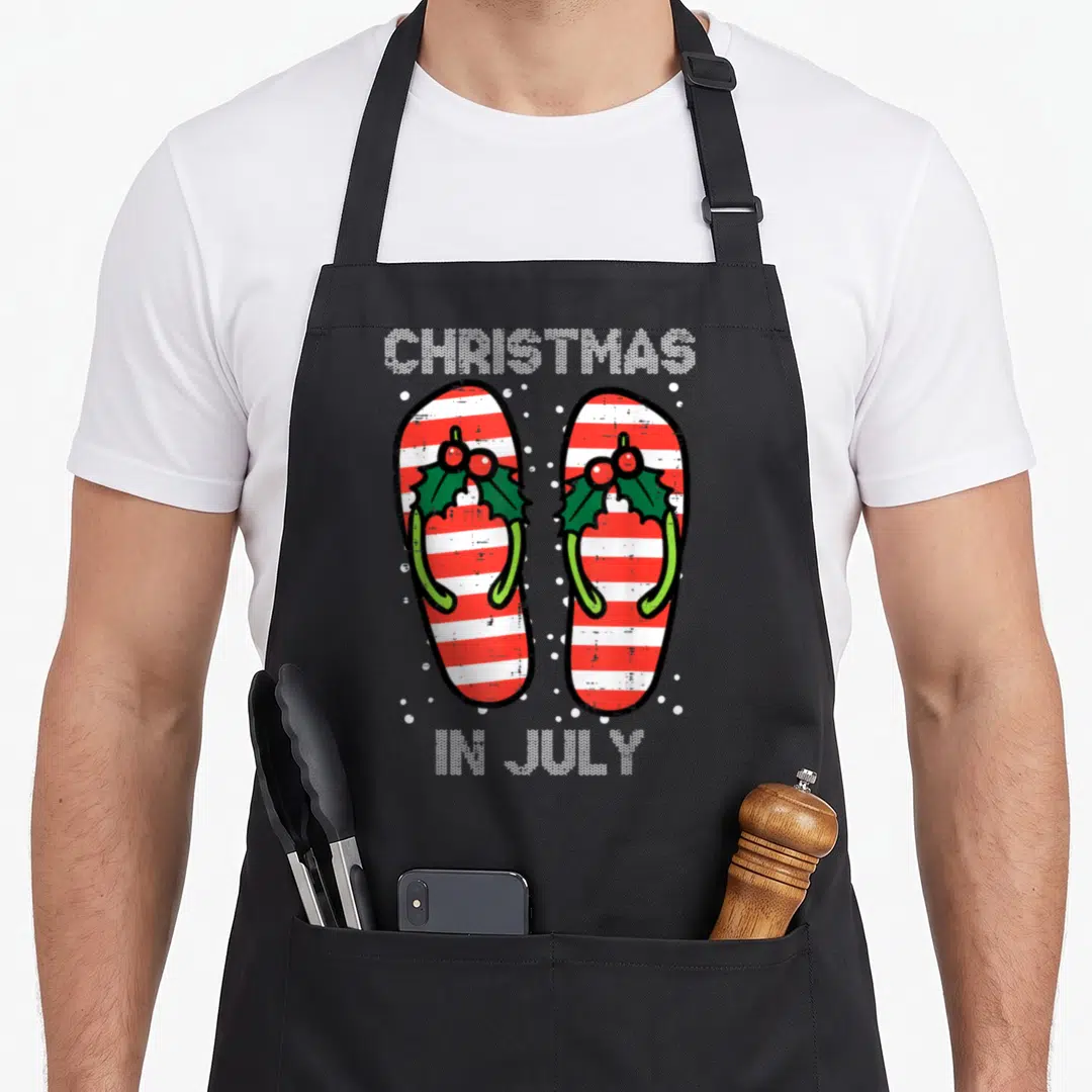 Flip christmas apron - Striped flip flops with holly and CHRISTMAS IN JULY text surrounded by white dots — shown on a black cotton canvas apron against a white background
