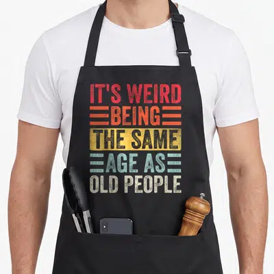 Birthday weird aprons - Stacked text IT'S WEIRD BEING THE SAME AGE AS OLD PEOPLE in red orange yellow teal — shown on a black cotton canvas apron against a white background