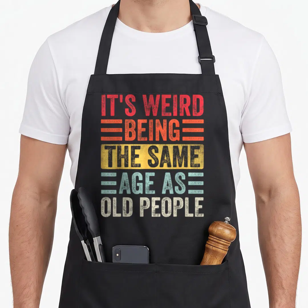 Birthday weird aprons - Stacked text IT'S WEIRD BEING THE SAME AGE AS OLD PEOPLE in red orange yellow teal — shown on a black cotton canvas apron against a white background