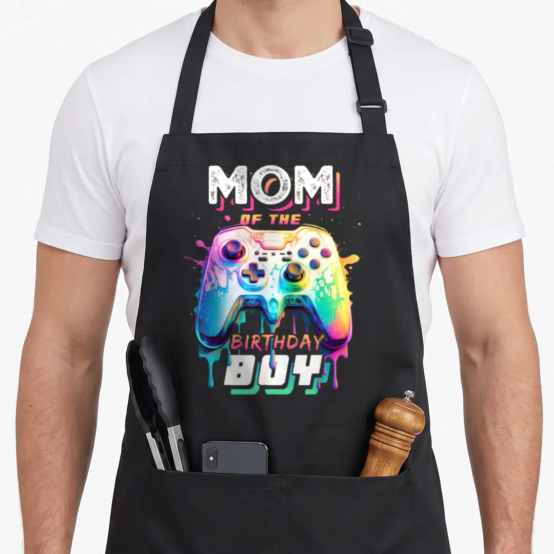 Cool birthday video game apron - Rainbow dripping game controller with Mom of the Birthday Boy text in white and orange — shown on a black cotton canvas apron against a white background