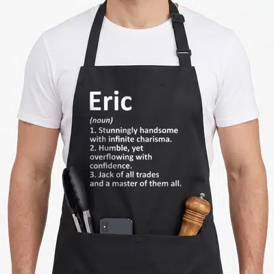 Eric father day aprons - White typography defining Eric as handsome and a master of all trades on grey background — shown on a black cotton canvas apron against a white background
