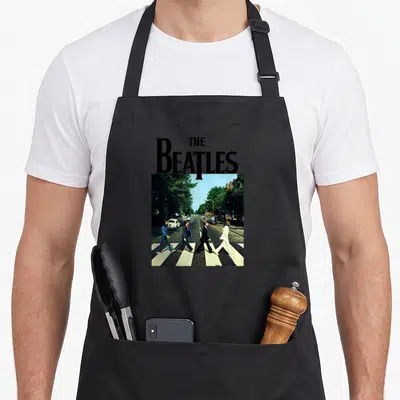Crossing abbey road apron - THE Beatles text above four men walking across a zebra crossing in a photo print — shown on a black cotton canvas apron against a white background