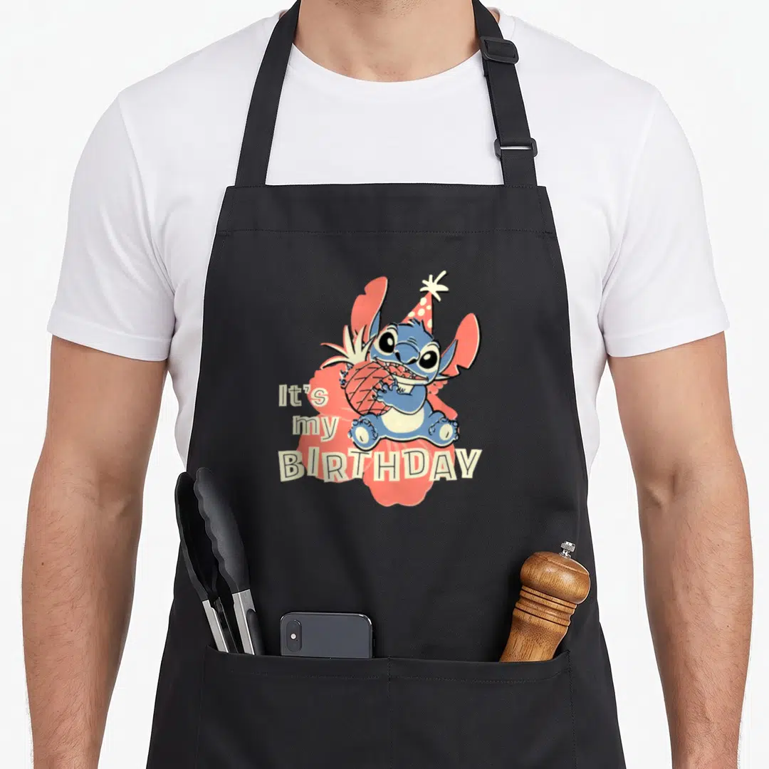 Birthday stitch apron - Blue cartoon character holding pineapple with It's My Birthday text on red splatter — shown on a black cotton canvas apron against a white background