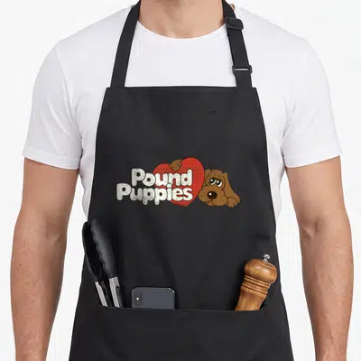 Pound cute puppy apron - White Pound Puppies text over red heart with brown cartoon dog peeking from behind — shown on a black cotton canvas apron against a white background