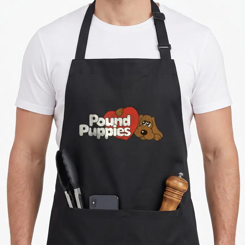 Pound cute puppy apron - White Pound Puppies text over red heart with brown cartoon dog peeking from behind — shown on a black cotton canvas apron against a white background