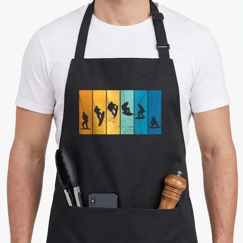 Vintage wakeboard wakeboarding apron - Six grey wakeboarder silhouettes arranged in orange, yellow, and blue vertical — shown on a black cotton canvas apron against a white background