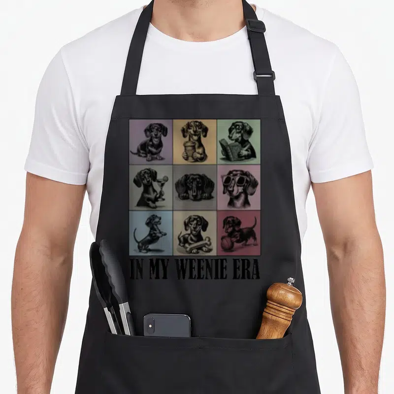 Dachshund era dog apron - Grid of nine cartoon dachshunds in colored squares above IN MY WEENIE ERA text — shown on a black cotton canvas apron against a white background