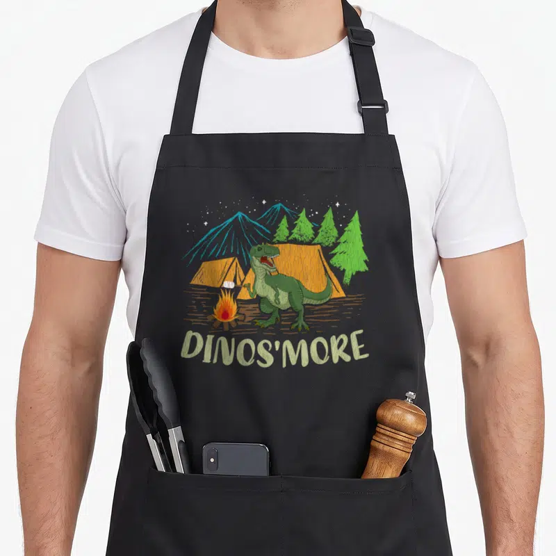Camping dinosaur apron - Green dinosaur roasting marshmallow over campfire with orange tents and blue mountains — shown on a black cotton canvas apron against a white background