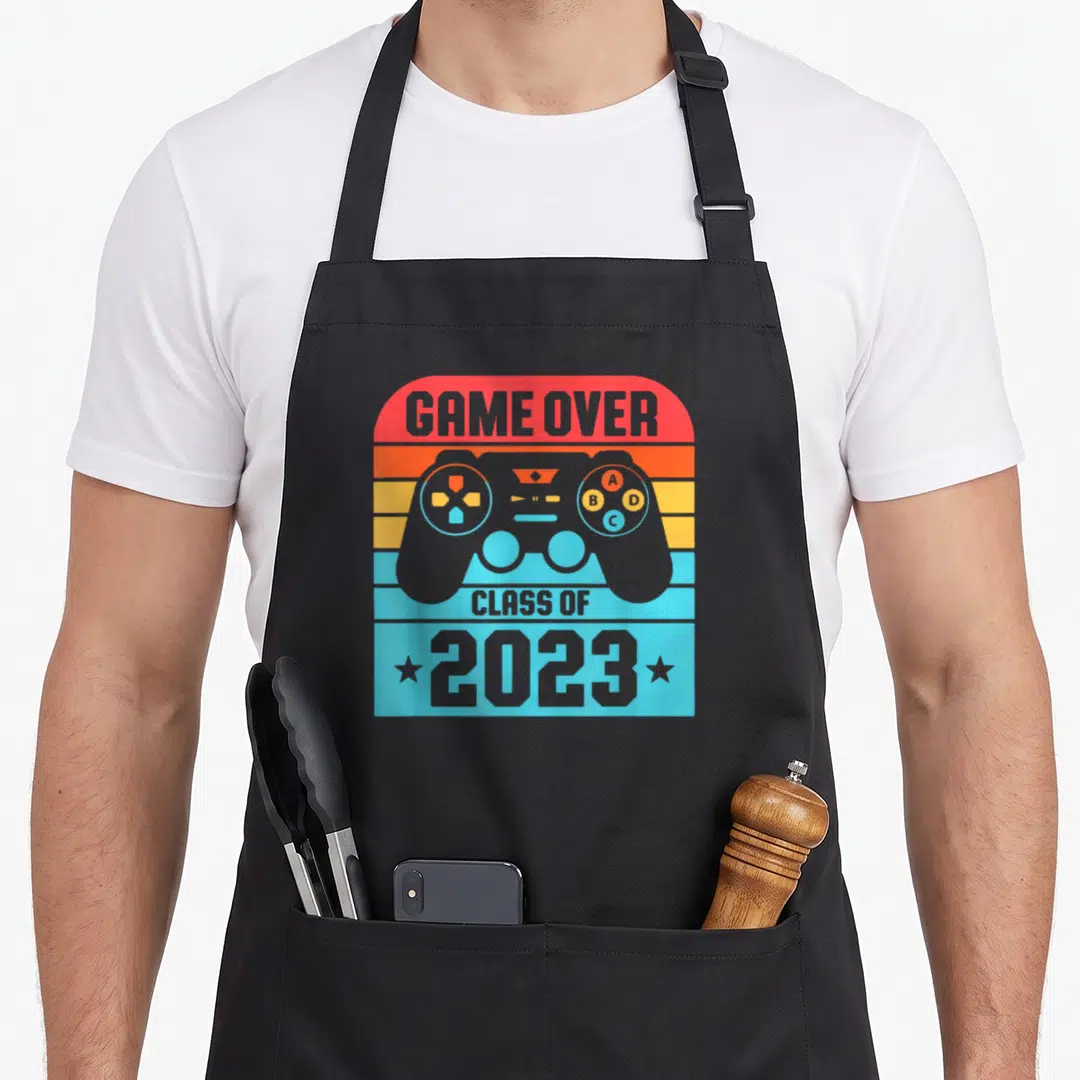 Class game aprons - Retro grey game controller with GAME OVER and Class of 2023 text on colorful stripes — shown on a black cotton canvas apron against a white background