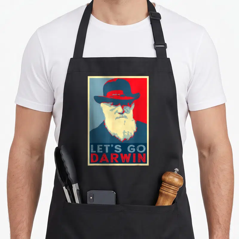 Darwin trendy apron - Stylized Darwin portrait with Let's Go Darwin text on split blue and red background — shown on a black cotton canvas apron against a white background