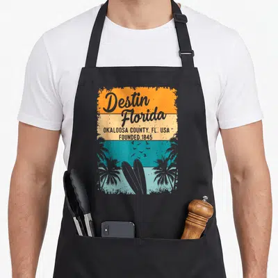 Destin beach florida apron - Retro striped graphic with Destin Florida text, two surfboards, palm trees, and birds — shown on a black cotton canvas apron against a white background