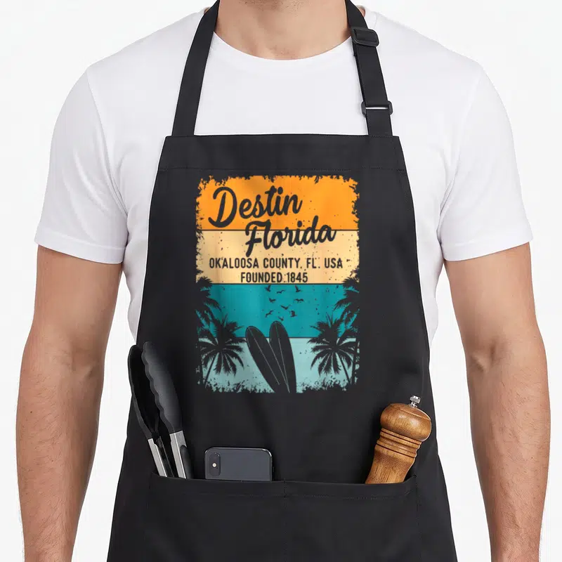Destin beach florida apron - Retro striped graphic with Destin Florida text, two surfboards, palm trees, and birds — shown on a black cotton canvas apron against a white background