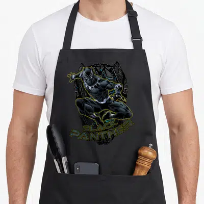 Trimmed black apron - Leaping Black Panther character outlined in yellow with text over a patterned black shield shape — shown on a black cotton canvas apron against a white background