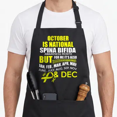 Support spina bifida awareness apron - Yellow and white text listing all months with two awareness ribbons at bottom — shown on a black cotton canvas apron against a white background