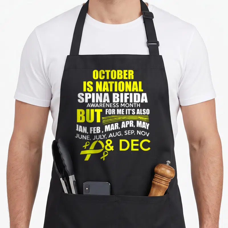Support spina bifida awareness apron - Yellow and white text listing all months with two awareness ribbons at bottom — shown on a black cotton canvas apron against a white background