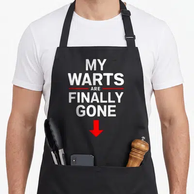 Trash wart apron - White text MY WARTS ARE FINALLY GONE with red lines and downward arrow on black background — shown on a black cotton canvas apron against a white background