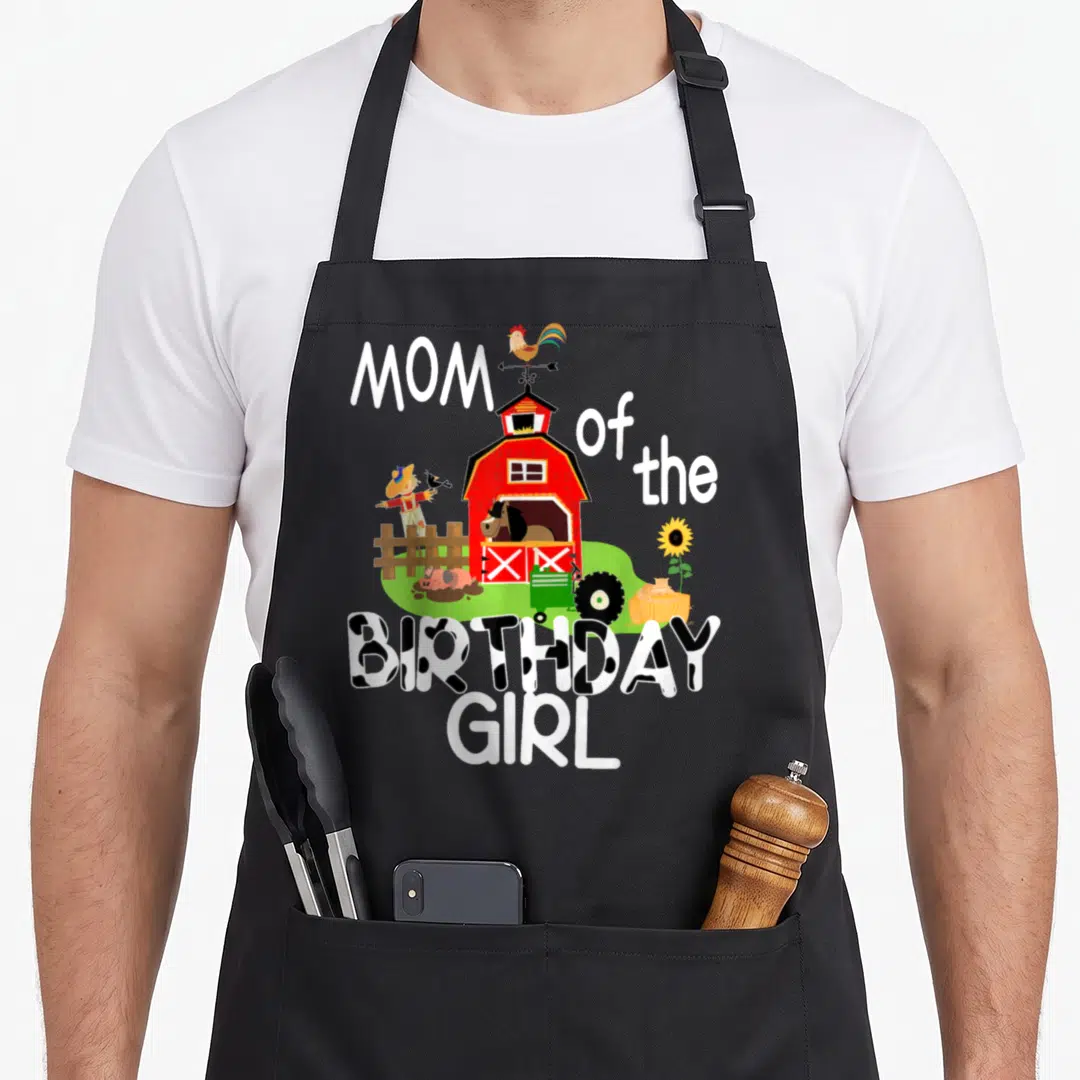 Barnyard cow print apron - Red barn with horse tractor and Mom of Birthday Girl cow print text — shown on a black cotton canvas apron against a white background