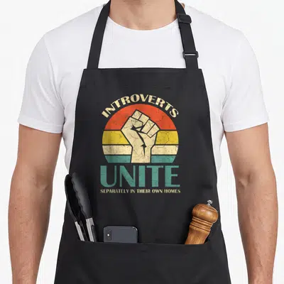 Unite introvert apron - Retro sunset with beige raised fist and arched text INTROVERTS UNITE SEPARATELY IN THEIR OWN — shown on a black cotton canvas apron against a white background