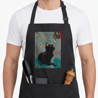 Blossom cat apron - Black cat illustration with cherry blossoms, teal leaves, and red kanji character inside brown — shown on a black cotton canvas apron against a white background