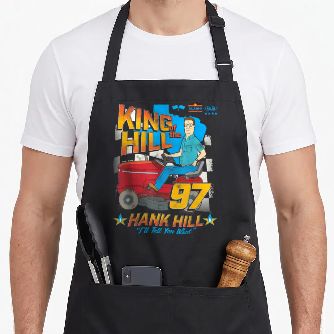 Hank cartoon apron - Hank Hill on red mower, KING of the HILL 97 text, checkered flag, I'll Tell You What quote — shown on a black cotton canvas apron against a white background