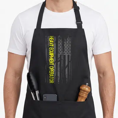 Machinery heavy canvas aprons - Vertical yellow HEAVY EQUIPMENT OPERATOR text beside a distressed grey USA flag pattern — shown on a black cotton canvas apron against a white background