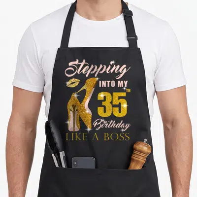 35th bbq boss apron - Gold glitter high heels and pink text reading Stepping Into My 35th Birthday Like A Boss — shown on a black cotton canvas apron against a white background