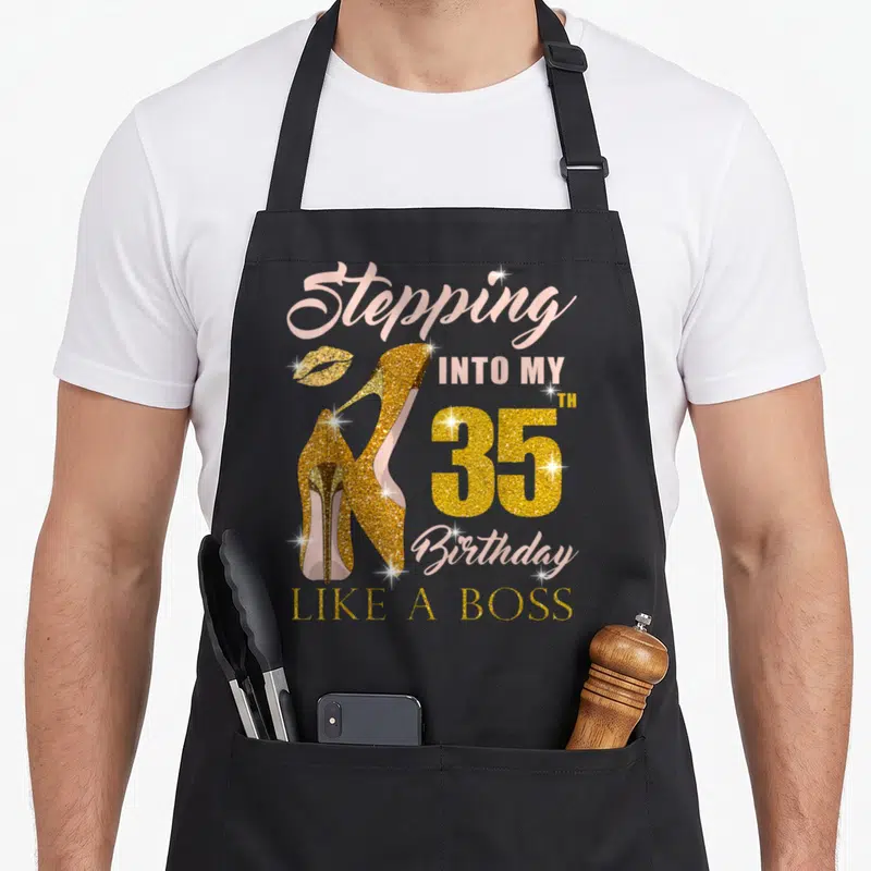 35th bbq boss apron - Gold glitter high heels and pink text reading Stepping Into My 35th Birthday Like A Boss — shown on a black cotton canvas apron against a white background