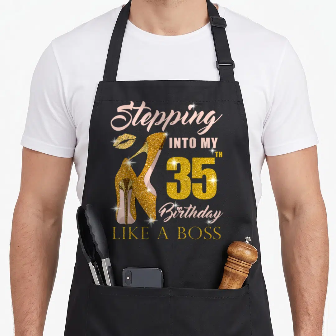 35th bbq boss apron - Gold glitter high heels and pink text reading Stepping Into My 35th Birthday Like A Boss — shown on a black cotton canvas apron against a white background