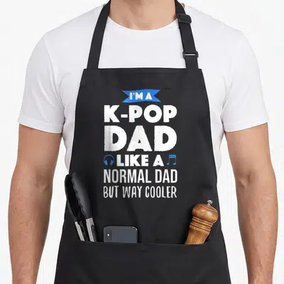 Pop music korean apron - Stacked white text I'M A K-POP DAD with blue banner, headphones, and musical note icon — shown on a black cotton canvas apron against a white background