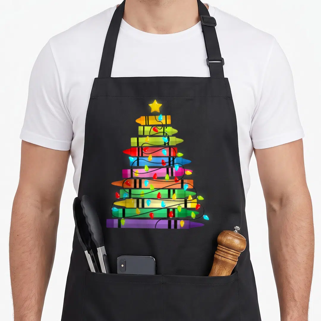 Crayon christmas tree apron - Stacked yellow green red blue purple crayons forming a tree with a star and lights — shown on a black cotton canvas apron against a white background