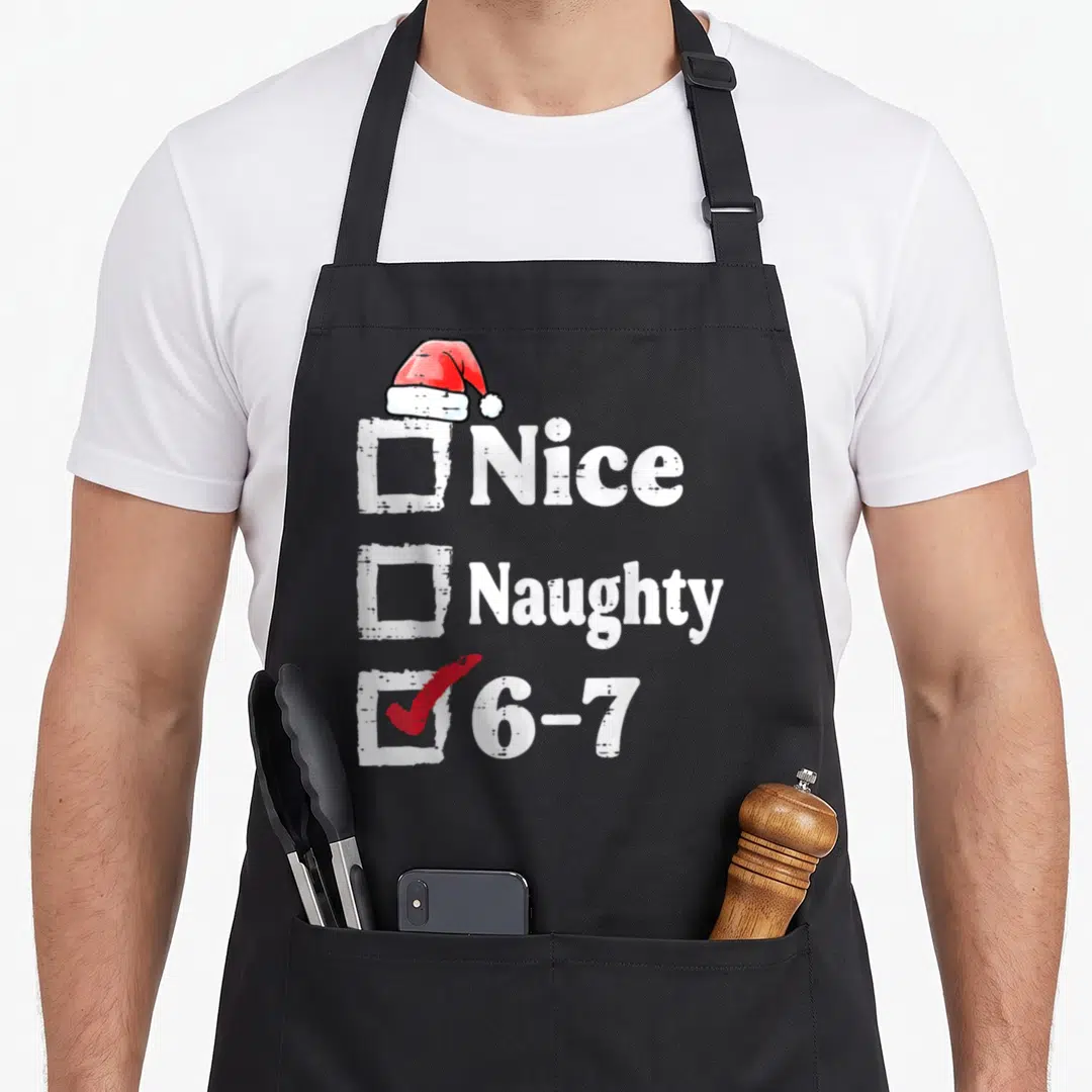 Meme christmas apron - Checklist with Santa hat checking option 6-7 next to nice and naughty labels — shown on a black cotton canvas apron against a white background
