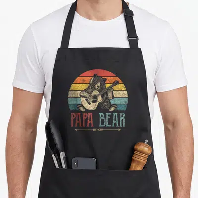 Guitar father day apron - Retro bear playing guitar in front of striped sun with PAPA BEAR text — shown on a black cotton canvas apron against a white background