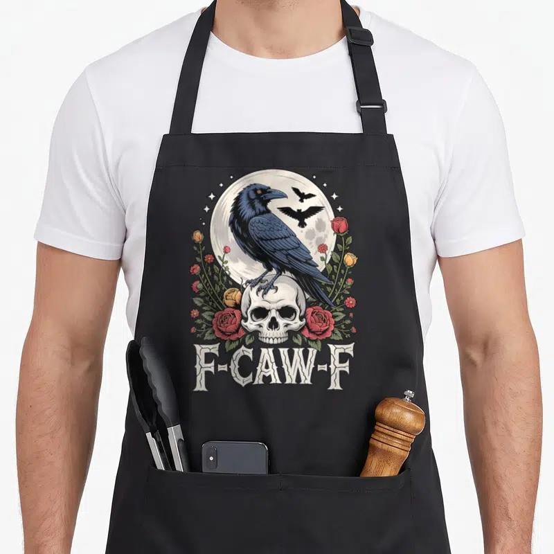 Cawing bird aprons - Dark blue raven on skull with red roses and F-Caw-F text — shown on a black cotton canvas apron against a white background