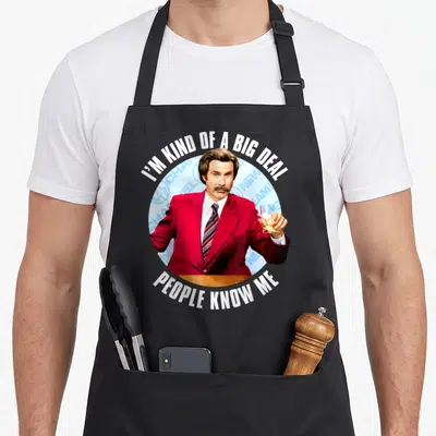 Anchorman lover comfort apron - Man in red suit holding amber drink circle with I'M KIND OF A BIG DEAL PEOPLE KNOW ME — shown on a black cotton canvas apron against a white background