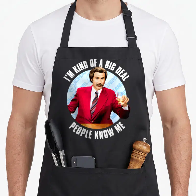 Anchorman lover comfort apron - Man in red suit holding amber drink circle with I'M KIND OF A BIG DEAL PEOPLE KNOW ME — shown on a black cotton canvas apron against a white background