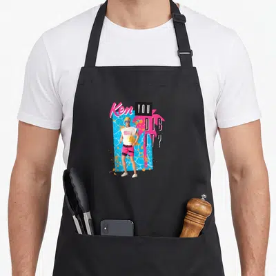 Dig general apron - Cartoon Ken doll holding yellow board, blue water, pink palm tree, Ken YOU DIG IT text — shown on a black cotton canvas apron against a white background