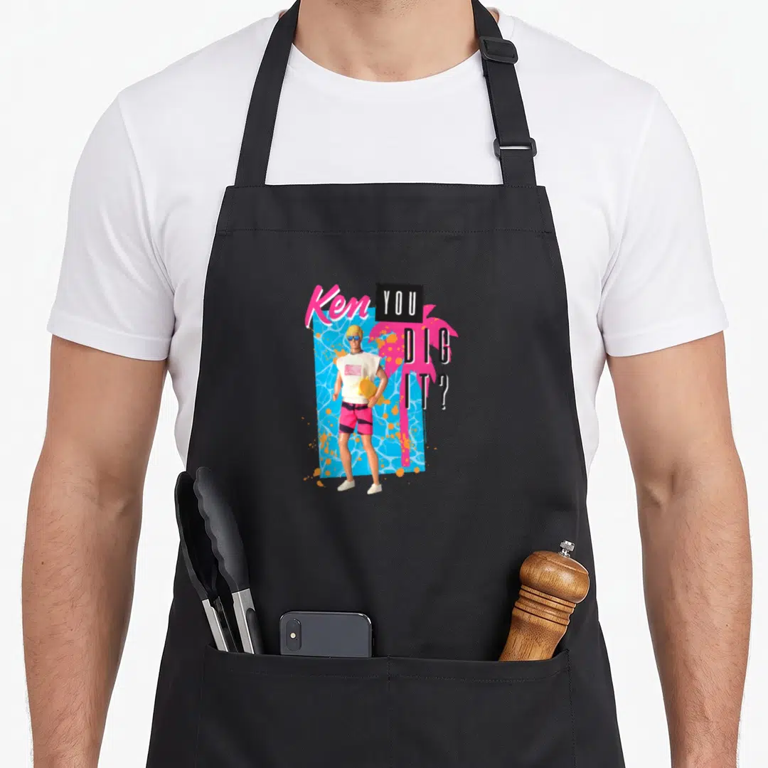Dig general apron - Cartoon Ken doll holding yellow board, blue water, pink palm tree, Ken YOU DIG IT text — shown on a black cotton canvas apron against a white background