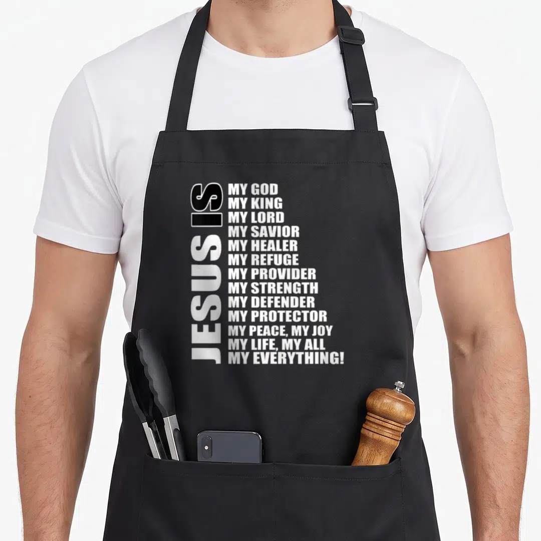 Phrase christian apron - White typography listing 13 titles of Jesus arranged vertically and horizontally on a black — shown on a black cotton canvas apron against a white background