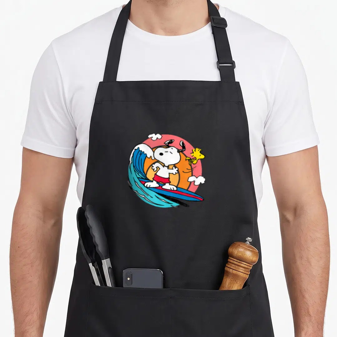 Surf decade apron - Snoopy surfing on blue wave with yellow Woodstock pink sun white clouds cartoon — shown on a black cotton canvas apron against a white background