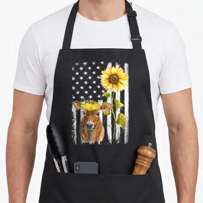 Cow sunflower apron - Watercolor brown cow with flower crown and yellow sunflower against distressed white flag stripes — shown on a black cotton canvas apron against a white background