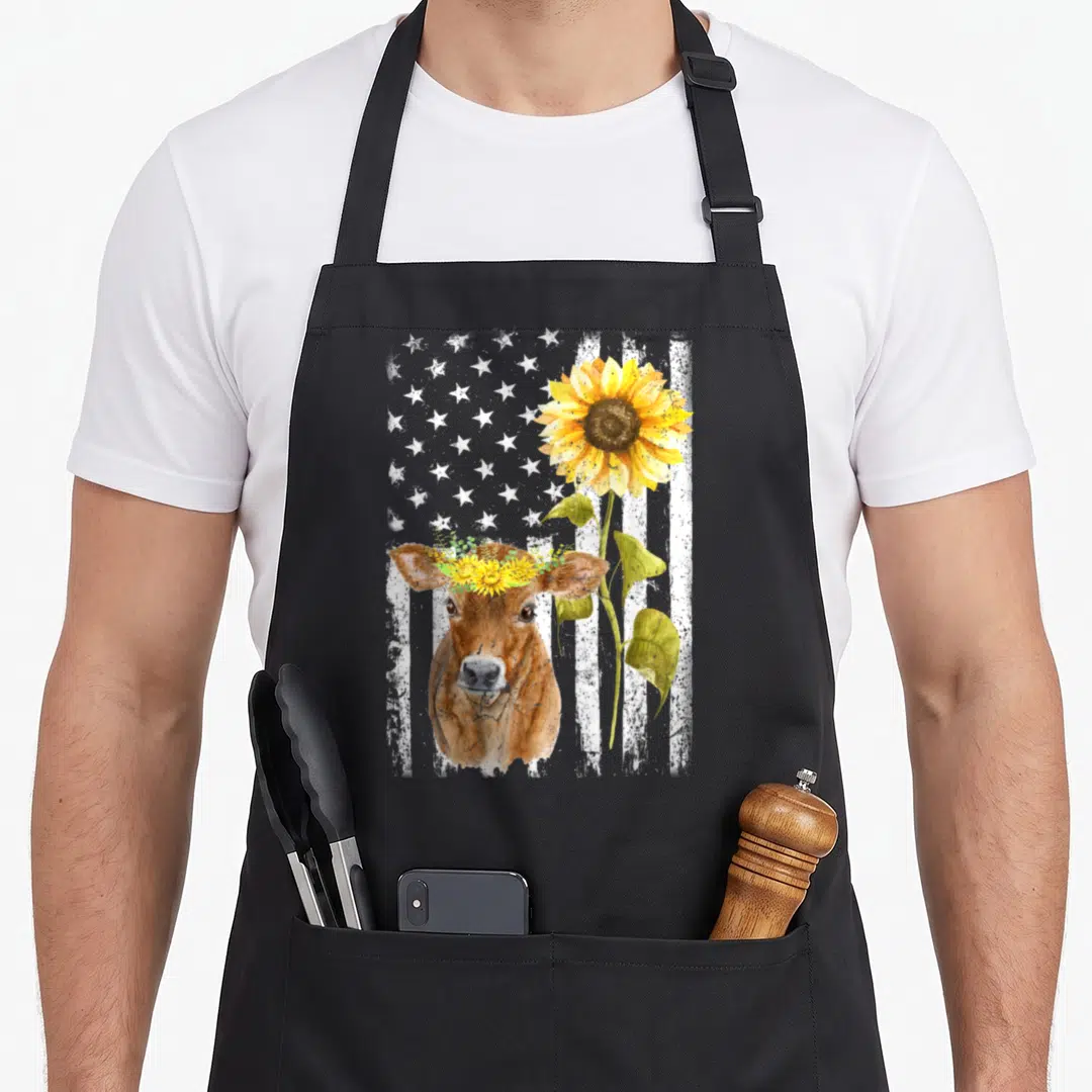 Cow sunflower apron - Watercolor brown cow with flower crown and yellow sunflower against distressed white flag stripes — shown on a black cotton canvas apron against a white background