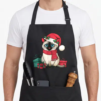 Xmas cute cat aprons - Cartoon Siamese cat in red Santa hat and scarf sitting among red and teal gift boxes — shown on a black cotton canvas apron against a white background