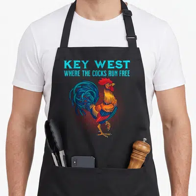 Run florida apron - Blue text reading Key West Where The Cocks Run Free above a colorful rooster on a red halftone — shown on a black cotton canvas apron against a white background