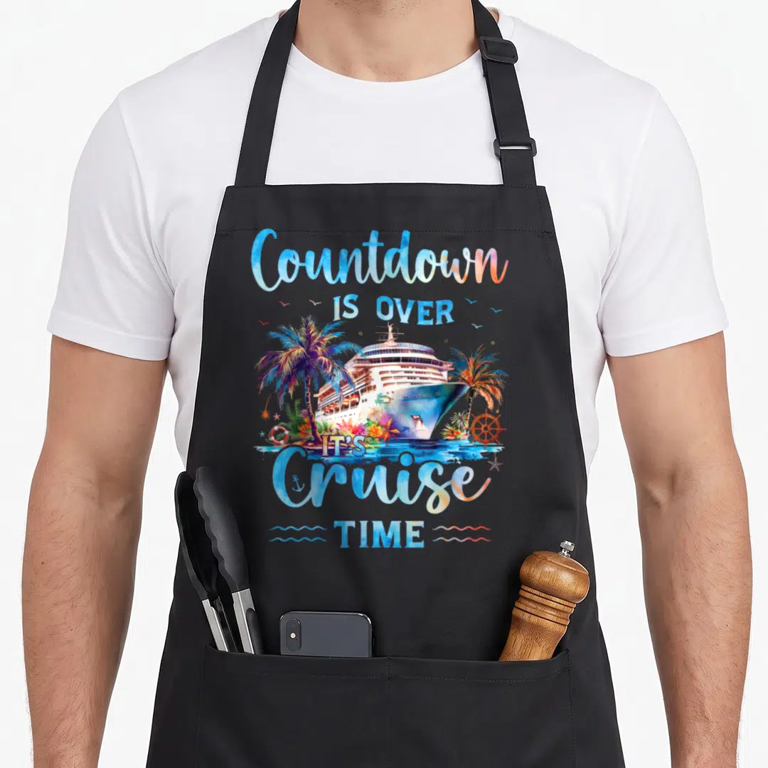 Countdown crazy cruise apron - Watercolor cruise ship flanked by palms with stacked blue and orange text reading — shown on a black cotton canvas apron against a white background