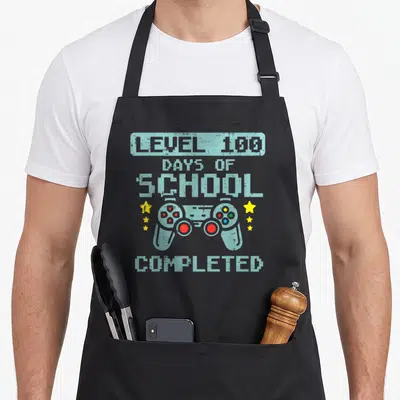 Completed game aprons - Pixel art controller with Level 100 Days of School Completed text and six yellow stars — shown on a black cotton canvas apron against a white background