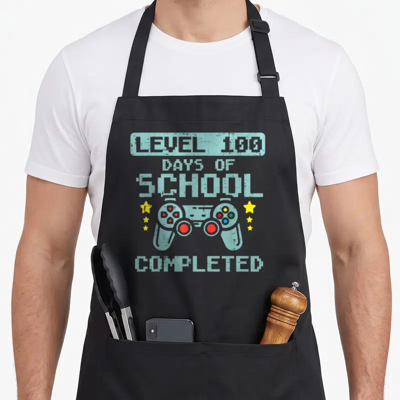 Completed game aprons - Pixel art controller with Level 100 Days of School Completed text and six yellow stars — shown on a black cotton canvas apron against a white background