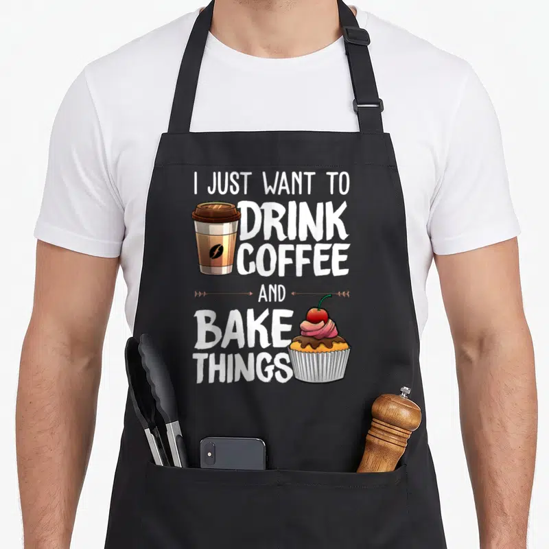 Drinking baker apron - Stacked text reading I Just Want to Drink Coffee and Bake Things with a coffee cup and cupcake — shown on a black cotton canvas apron against a white background