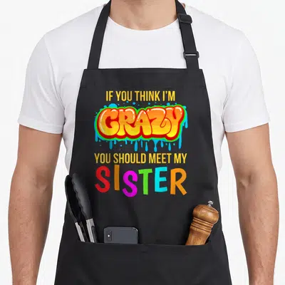 Meet novelty apron - Stacked colorful text reading If You Think I'm Crazy You Should Meet My Sister in yellow orange — shown on a black cotton canvas apron against a white background