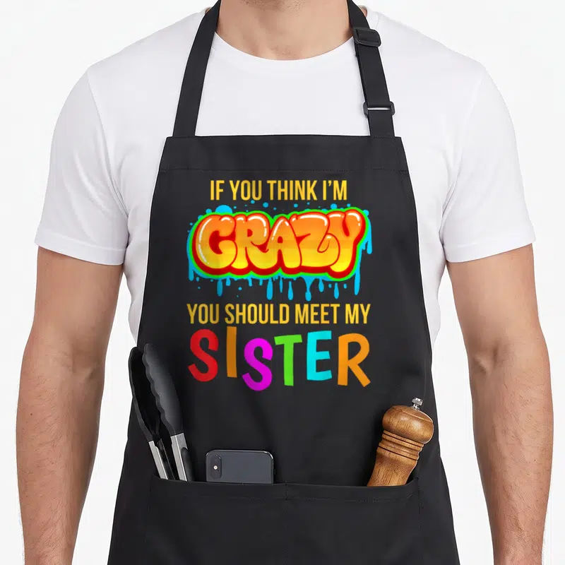 Meet novelty apron - Stacked colorful text reading If You Think I'm Crazy You Should Meet My Sister in yellow orange — shown on a black cotton canvas apron against a white background