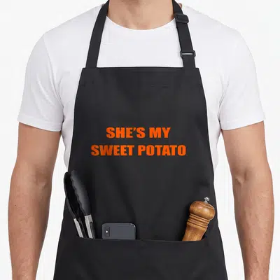 Goal potato apron - Orange text reading She's My Sweet Potato stacked in two lines — shown on a black cotton canvas apron against a white background